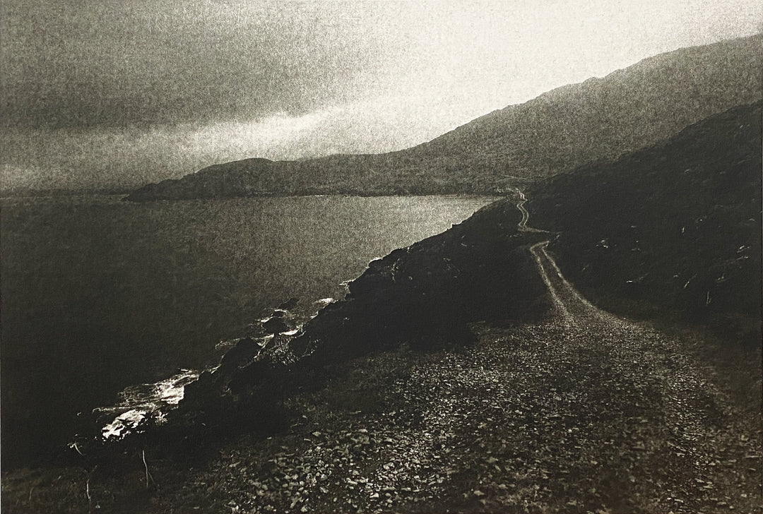 A clear road ahead is seen along the edge of a hill by the ocean. The sky above is bright. A silver gelatin photograph from a paper negative.