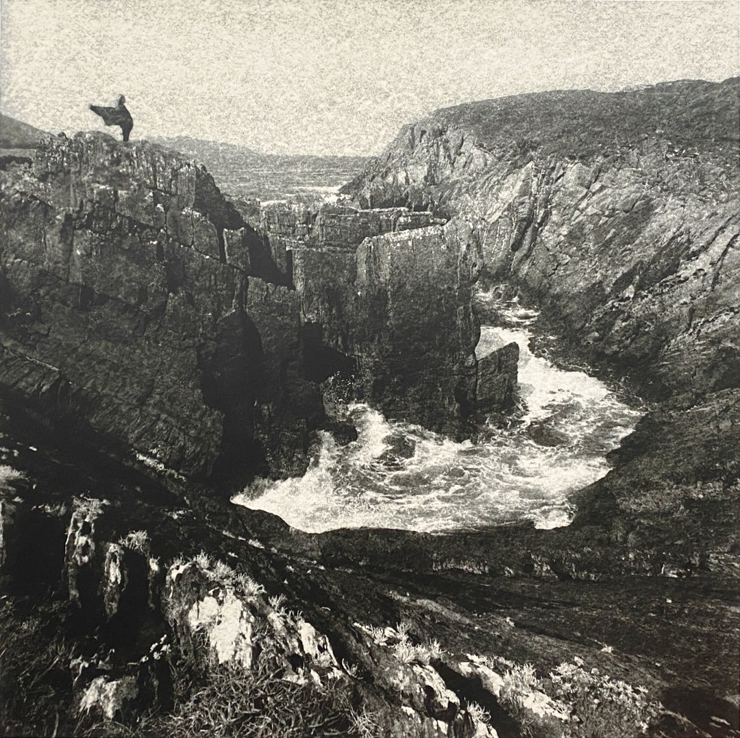 A lone figure stands on a rocky cliff above swirling waters  of the ocean. Silver gelatin photograph from a paper negative.